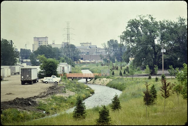Color photograph depicting developments along one side of a creek: on the left side of the photo, trucks and cars are parked on dirt. The right side is green and grassy with plenty of young spruce trees and larger oak trees in the background.